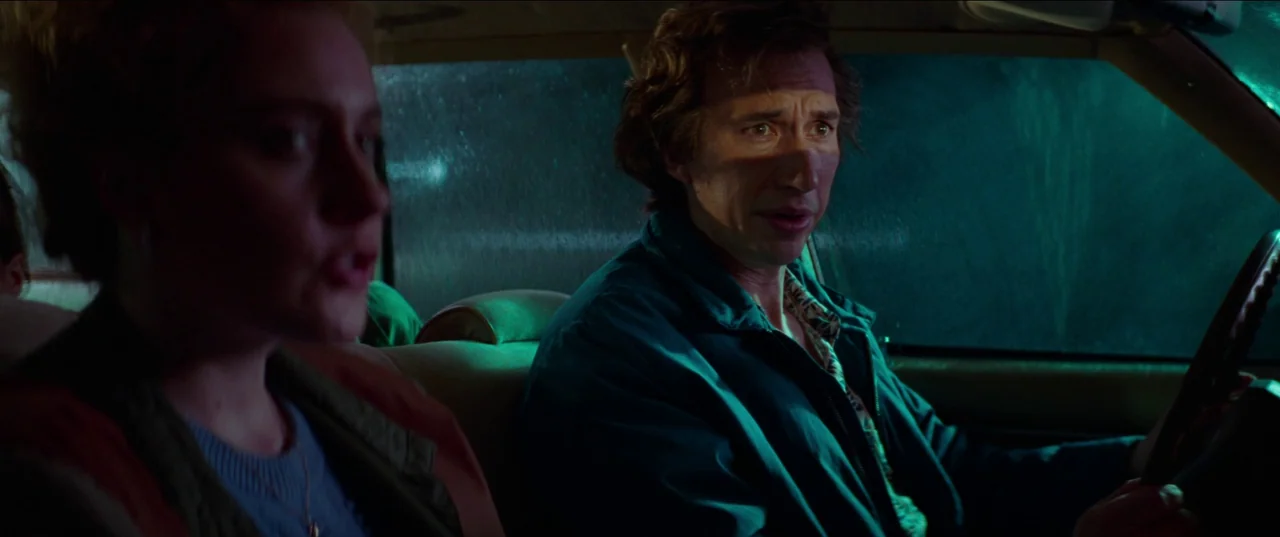 White Noise (2022) movie still: medium — The interior of a car at night illuminated by harsh green exterior light. A man drives while speaking to a…