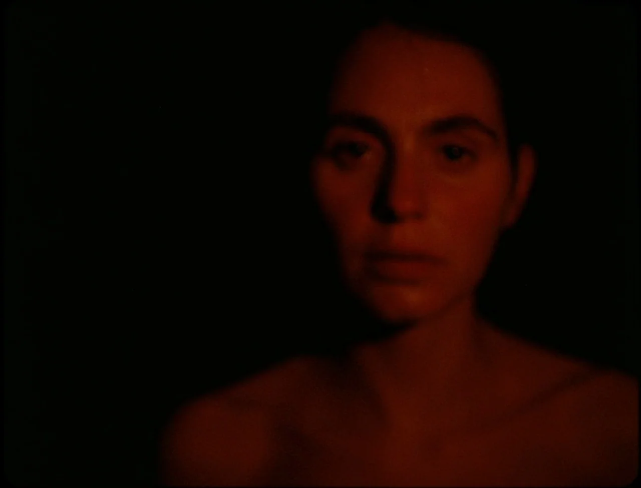 Godland (2022) movie still: closeup — A woman's face is partially lit in an otherwise pitch-black room. She is positioned frontally with her bare…
