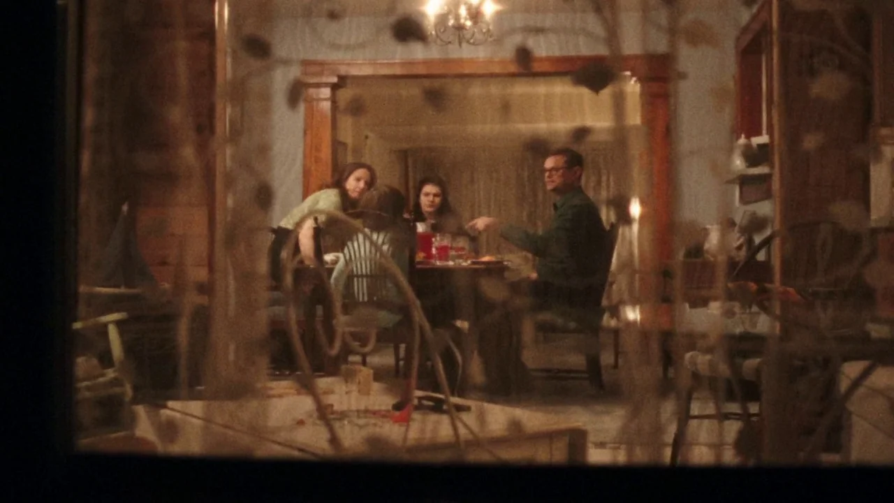 I Am Not a Serial Killer (2016) movie still: wide — A view through a lace curtain into a traditional wood-paneled residential dining area. A family…
