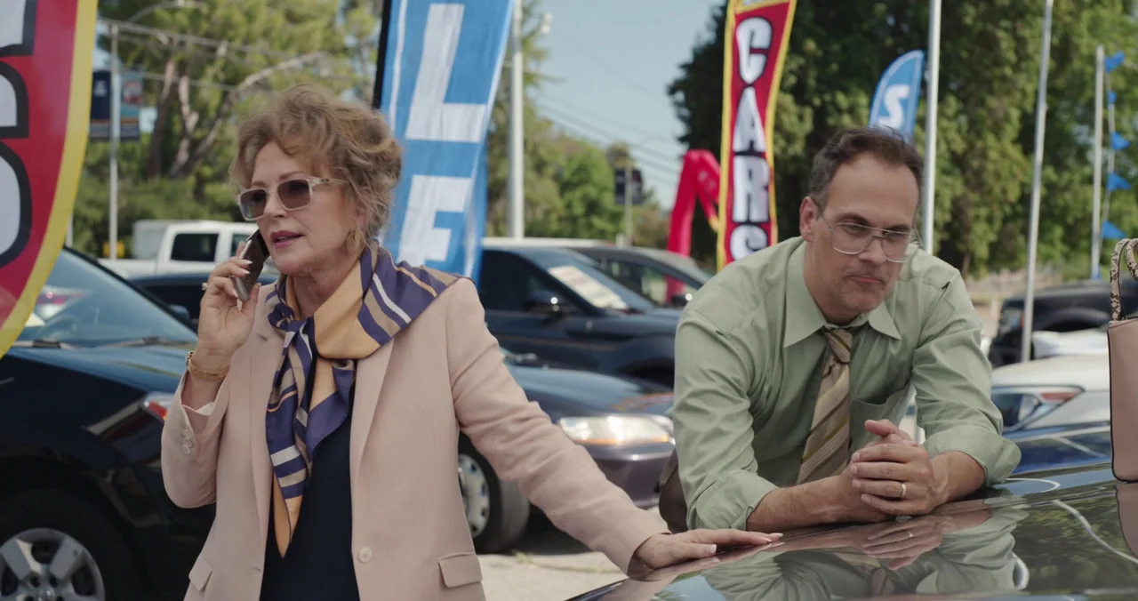Violet (2021) (2021) movie still: medium — A sunlit car dealership features rows of vehicles and colorful promotional pennant banners. An older…