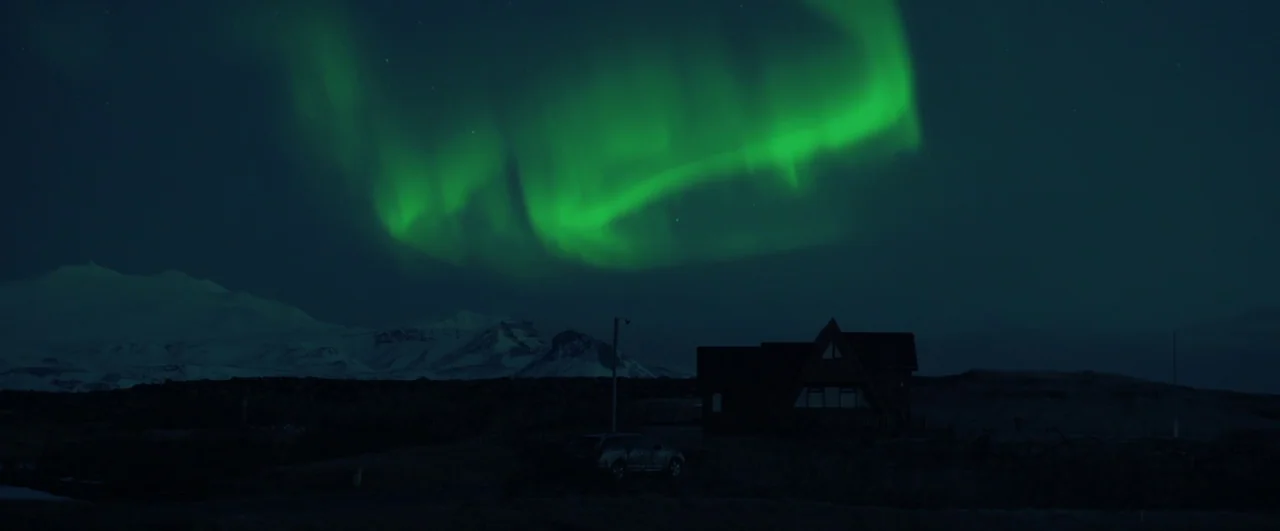 Rift (2017) movie still: establishing — A dark, snow-dusted landscape features a small house under a vast, dark sky. Intense green aurora borealis…