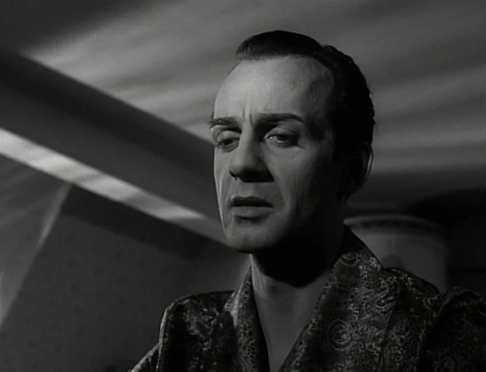 The Devil’s Eye (1960) movie still: closeup — A dimly lit room with stylized interior architectural features visible in the soft shadows. A man in a…