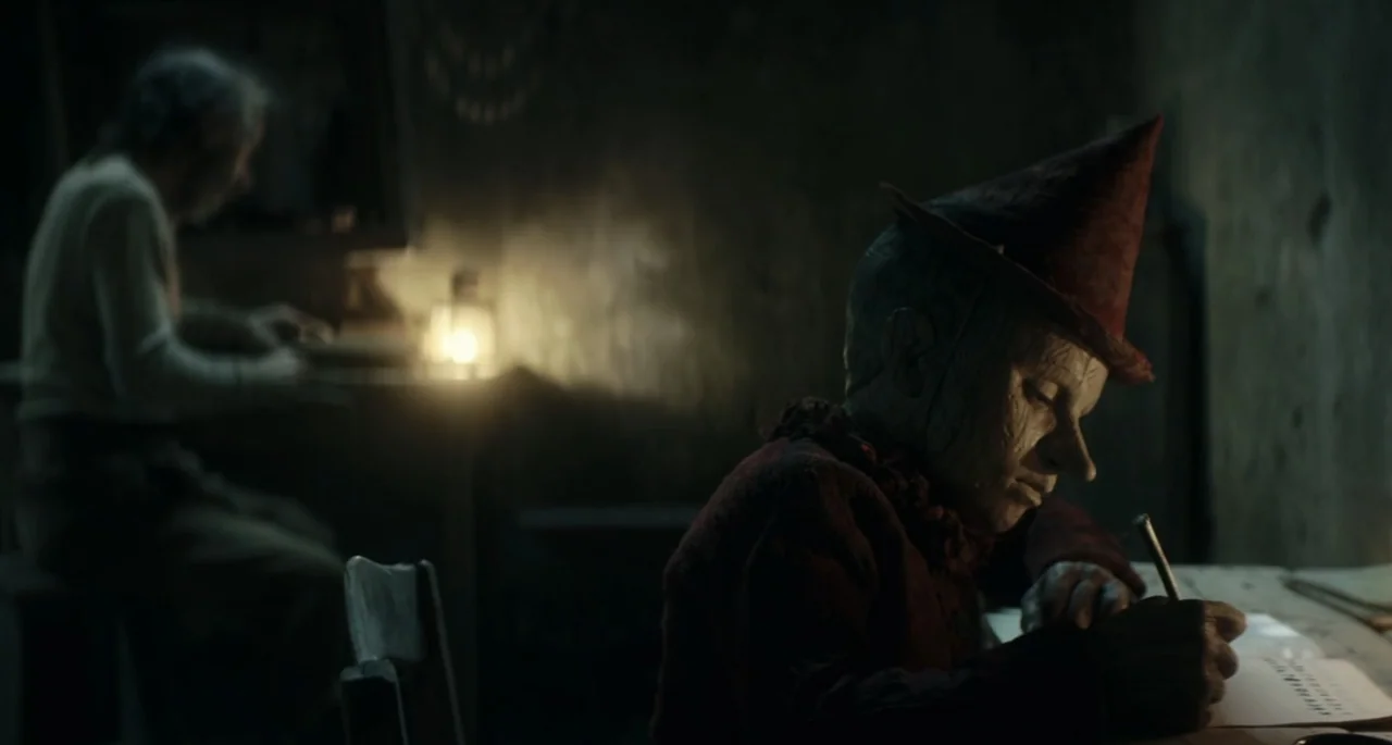 Pinocchio (2022) movie still: medium — A dimly lit room contains wooden furniture and surfaces worn from heavy use. A wooden puppet character sits…