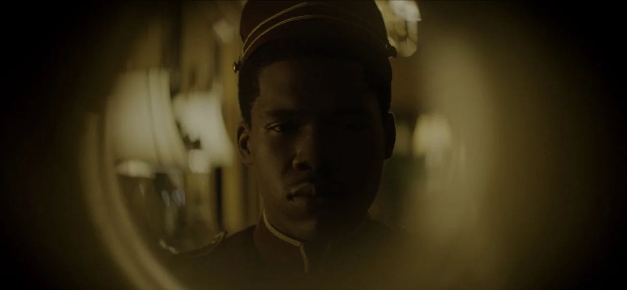 No Sudden Move (2021) movie still: closeup — A dimly lit hotel hallway with soft, blurry lamps in the background. A young man wearing a bellhop…