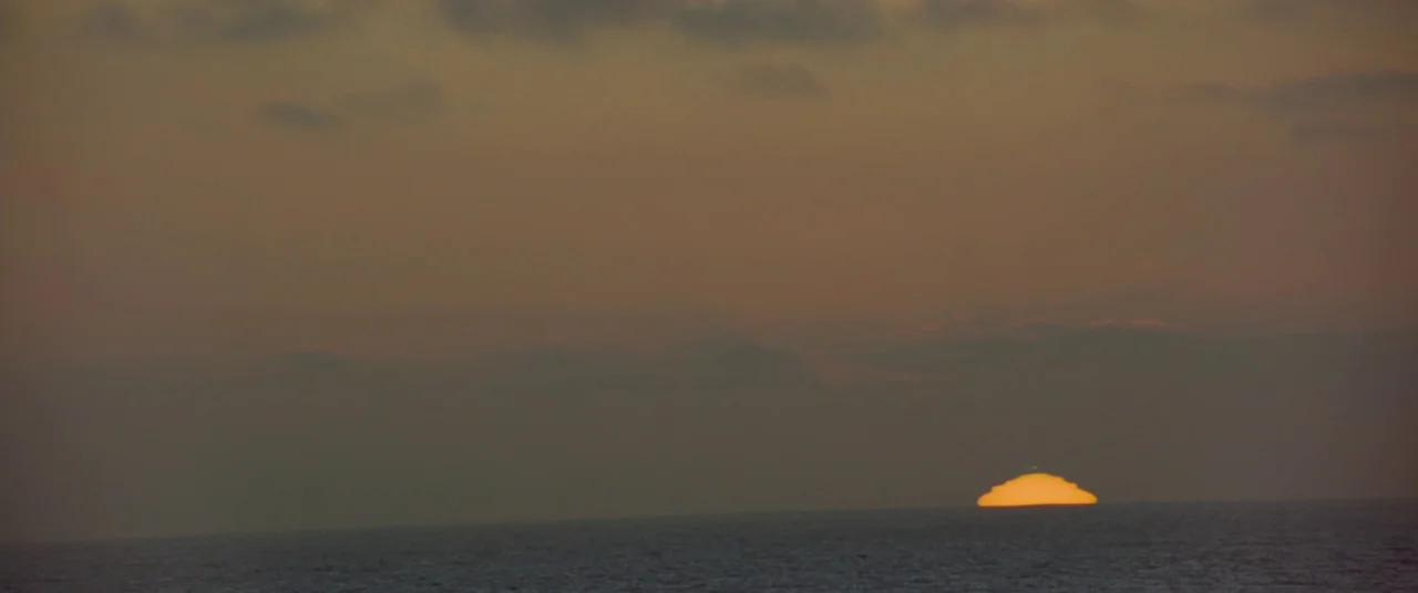 All Is Lost (2013) movie still: establishing — A wide ocean horizon during sunset with thick cloud cover obscuring the sky. The sun is nearly fully…