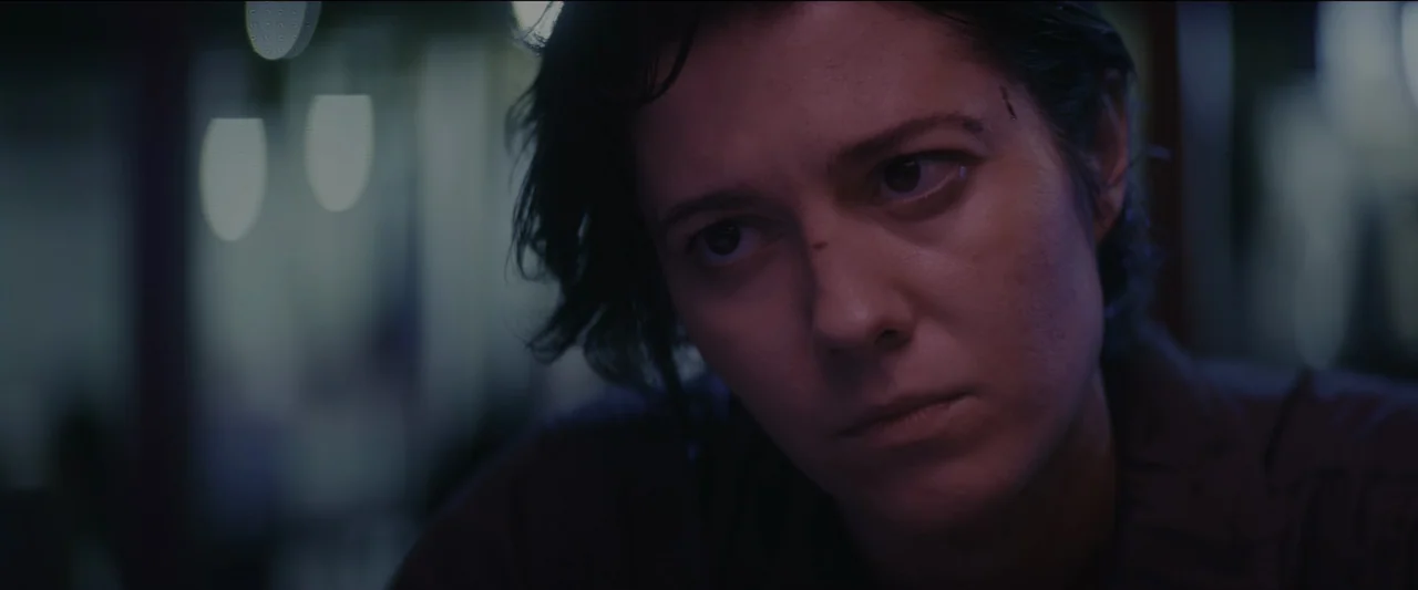 Kate (2021) movie still: closeup — The interior of a vehicle is dimly lit with cool, desaturated colors. A woman with short dark hair sits inside…