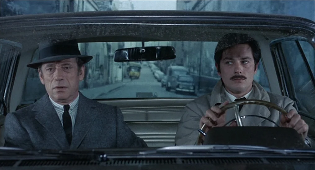 Le Cercle Rouge (1970) movie still: medium — A man and his driver sit in the front seat of a car in motion. The street behind them is visible…