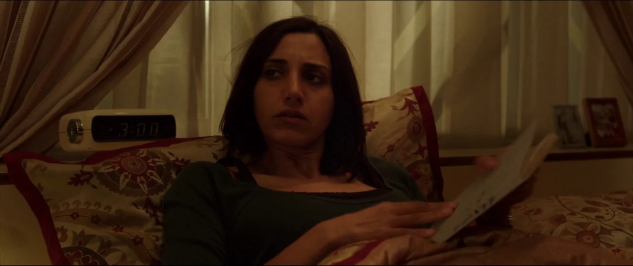 Under The Shadow (2016) movie still: medium — A woman lies in a bed with patterned pillows and curtains in the background. She holds an open book…