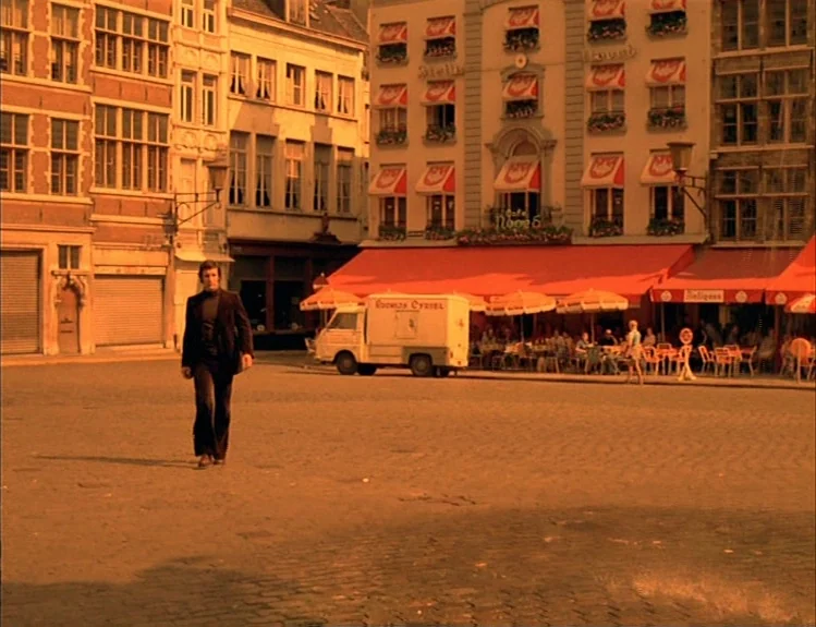 The Arrival of Joachim Stiller (1976) movie still: wide — A European town square features old architecture and a cafe with red awnings. A man walks…