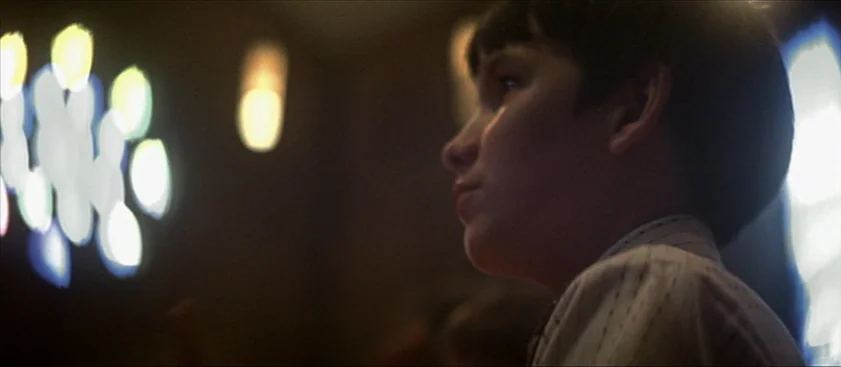 Nashville (1975) movie still: closeup — A dimly lit space features a large out-of-focus stained glass window on the side. A young boy looks toward…