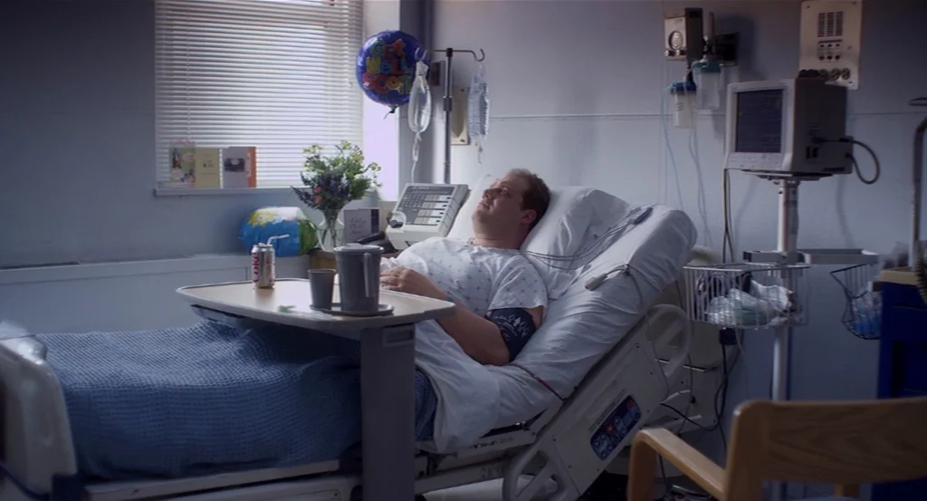 Dark Horse (2011) movie still: medium — A standard hospital room with white walls, a patient bed, and medical monitoring equipment. A man wearing a…