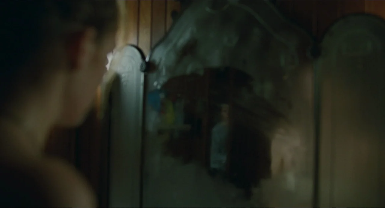 The Lodge (2019) movie still: closeup — A dimly lit room features a vintage wooden tri-fold vanity mirror with ornate edges. A person stands before…
