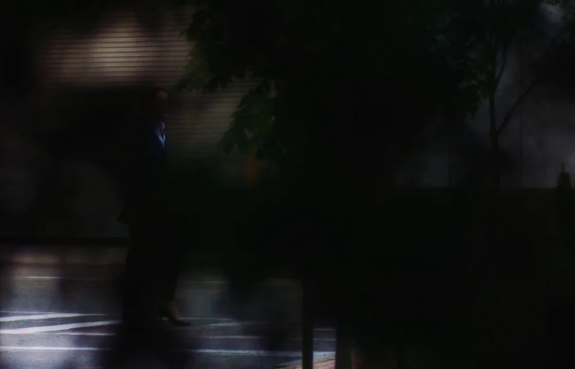 Unsane (2018) movie still: wide — A dark city street at night is framed through blurry foreground leaves and branches. A woman in a dark suit walks…