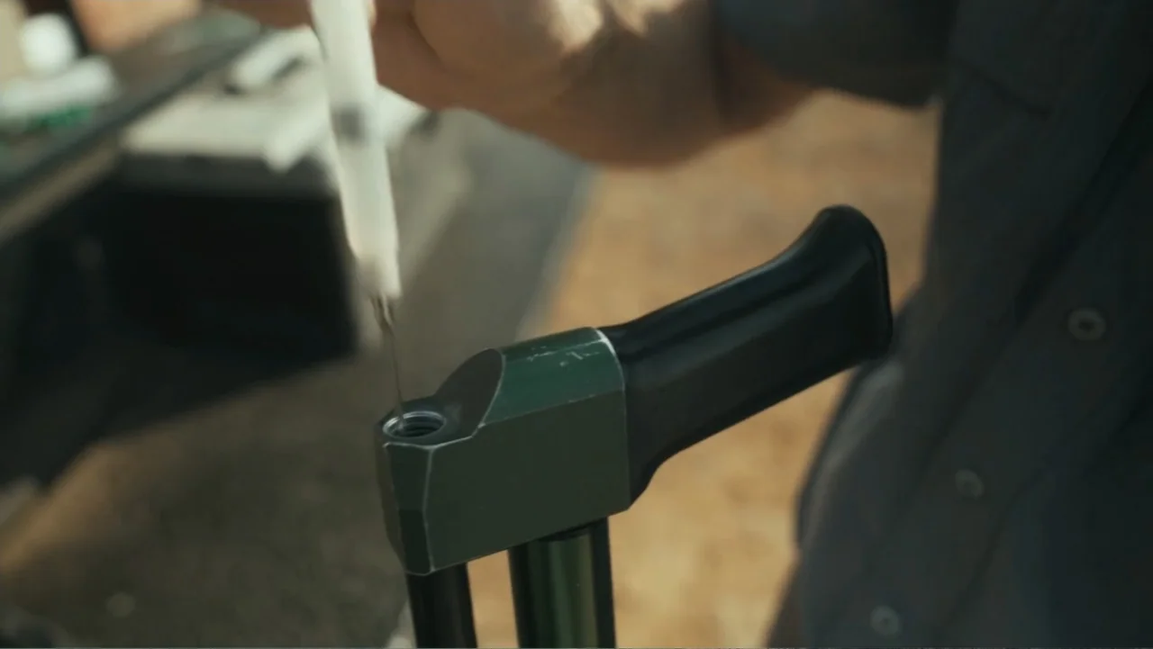 Bloody Trophy (2025) movie still: closeup — An outdoor workspace features a metal mechanical assembly viewed from a close perspective. A person…
