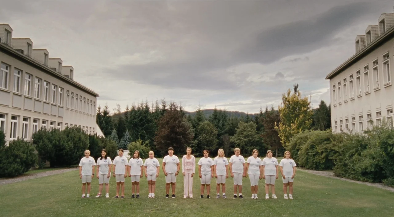 Paradise: Hope (2013) movie still: fullbody — A group of teenagers and one adult stand in a line on grass. Two institutional buildings face each…