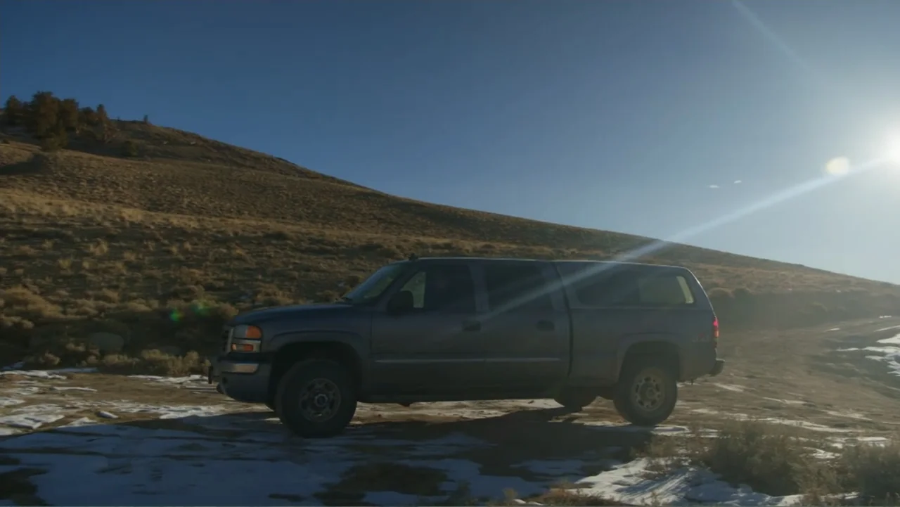 Trees and Other Entanglements (2023) movie still: wide — A dark grey pickup truck is parked on a dirt slope covered with patchy snow. The vehicle…