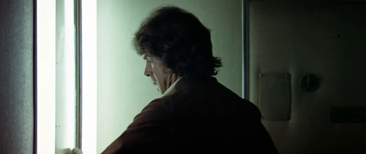 The Parallax View (1974) movie still: medium — A man stands in a dark corridor next to a glowing vertical light panel. He peers through a glass…