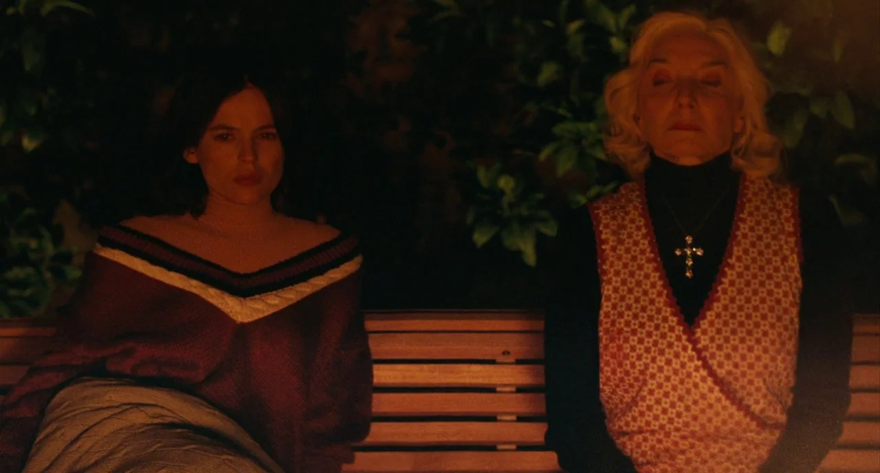 The Skin I Live In (2011) movie still: medium — Two women sit together on a wooden bench at night in front of dark foliage. The scene is illuminated…