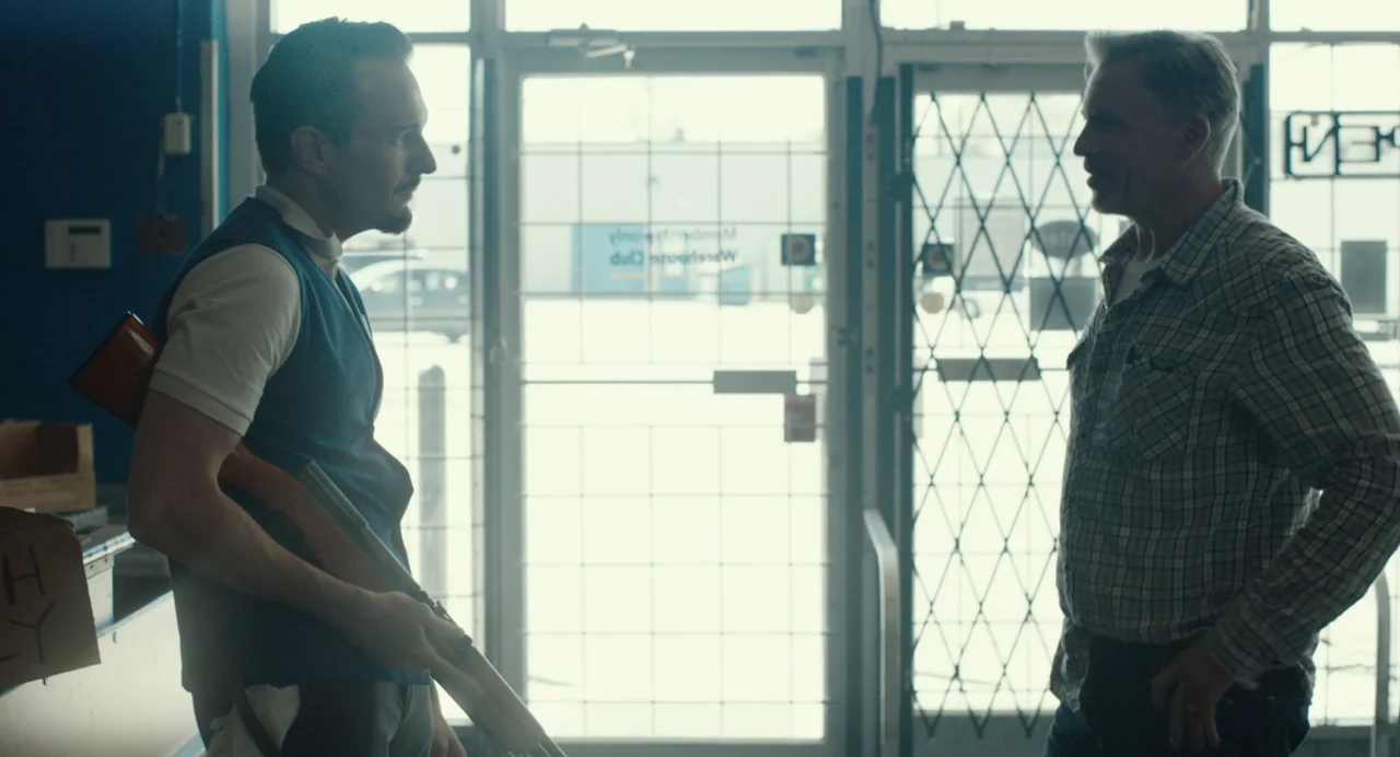 En lo profundo del bosque movie still: medium — A cluttered retail interior features a glass entrance door with a metal security gate. Two men stand…