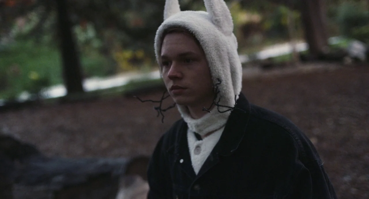 Palo Alto (2013) movie still: medium — An outdoor area with blurred trees and dark ground in the background. A young man wearing a white fleece hood…