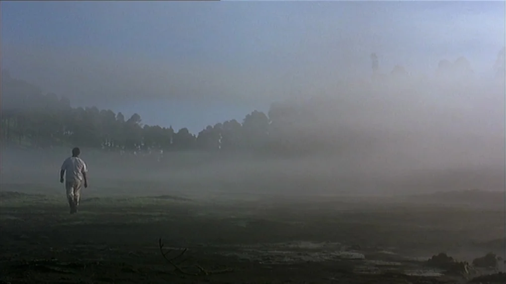 Battle In Heaven (2005) movie still: wide — A vast, damp field is enveloped in a thick layer of morning fog. A lone man walks steadily away from the…