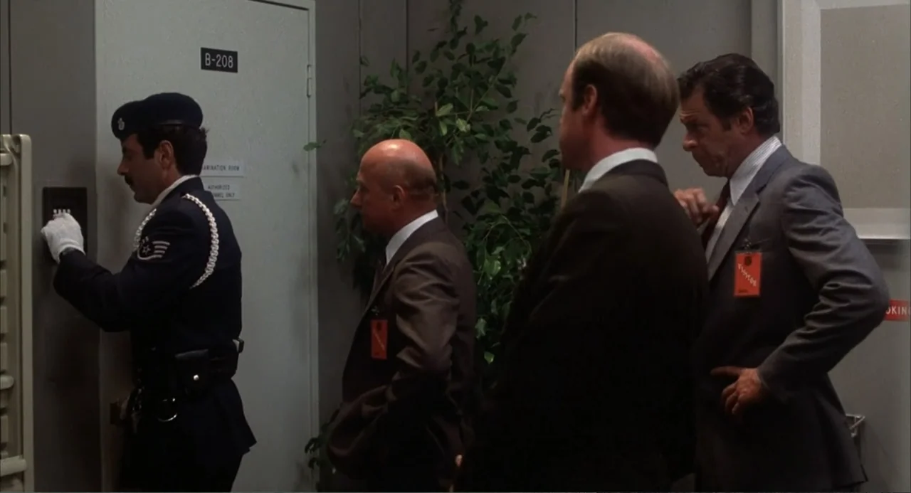 WarGames (1983) movie still: fullbody — A stark hallway with gray walls and a potted plant stands outside room B-208. A security guard presses a…
