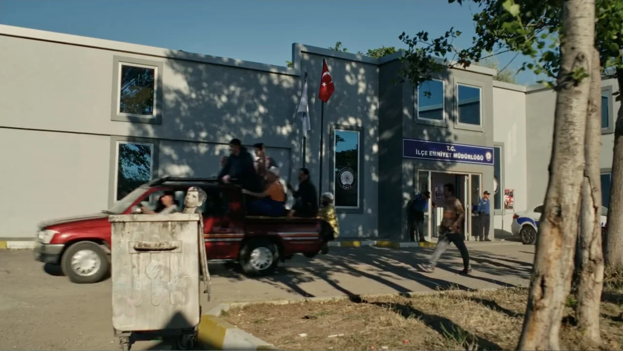 Paranormal Cuma (2024) movie still: wide — The exterior of a modern police building features grey concrete walls and flagpoles. A red pickup truck…
