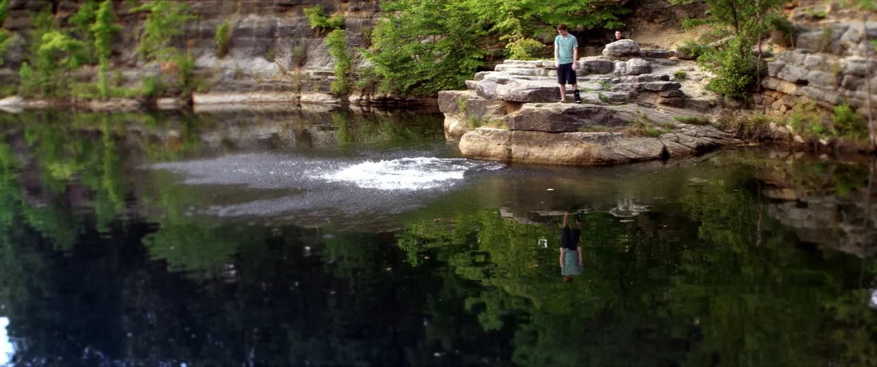 Kings Of Summer (2013) movie still: wide — A deep stone quarry with layered rocky walls and surrounding green trees. A teenager stands on the edge…