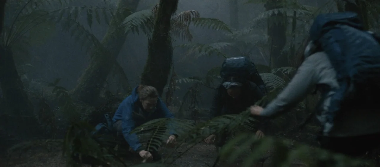 Force of Nature: The Dry 2 (2024) movie still: wide — A dense rainforest environment is filled with large ferns and dark mossy tree trunks. Three…