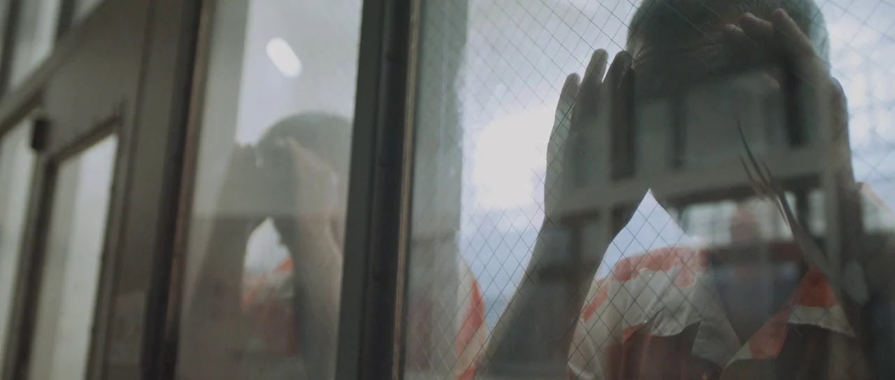 To The Wonder (2012) movie still: medium — A visitation room with a thick glass partition embedded with a wire mesh grid. A man wearing an orange…