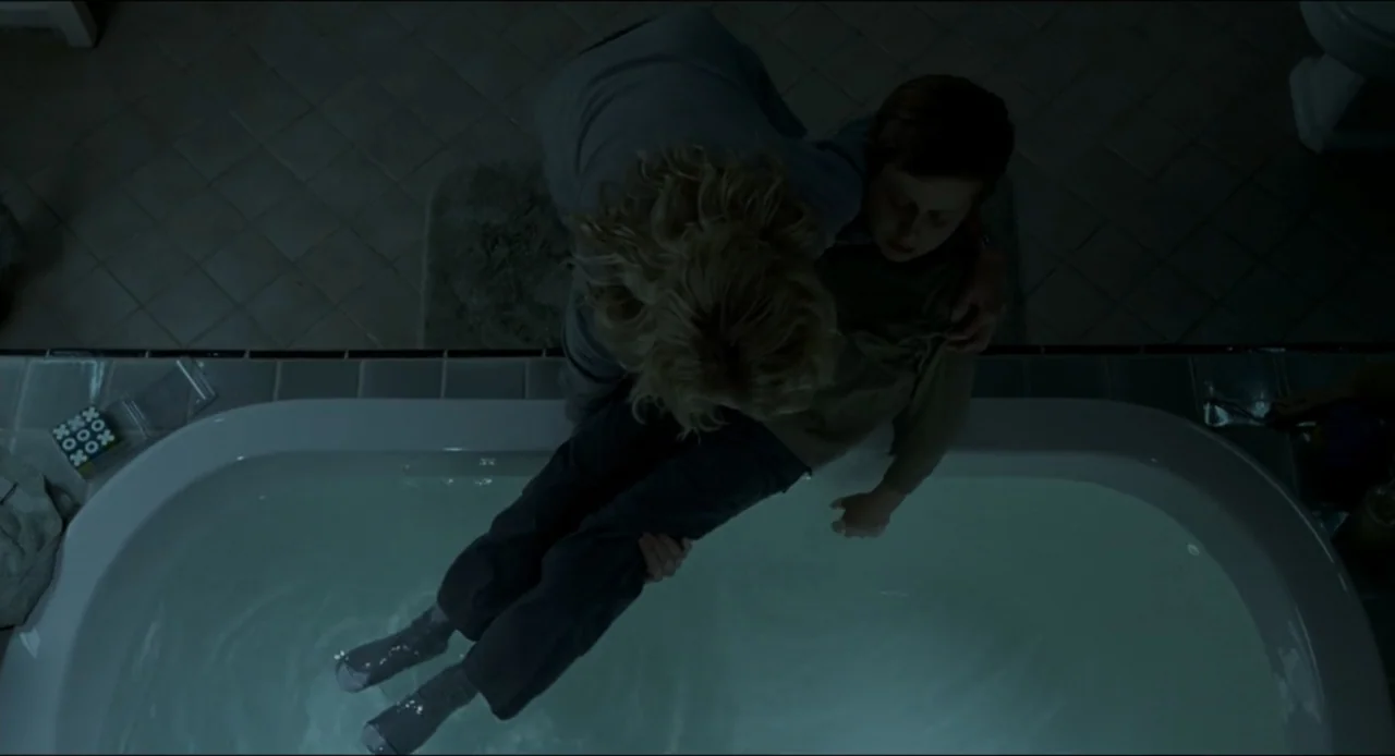 The Ring Two (2005) movie still: medium — A dimly lit tiled bathroom features a large bathtub filled with water. A woman leans over the tub to pull…