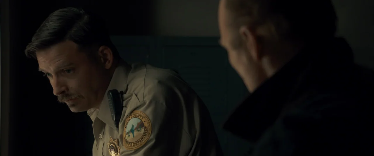 Frontera (2014) movie still: medium — A dark office space features lockers in the background near a desk. A sheriff in uniform sits at a desk…