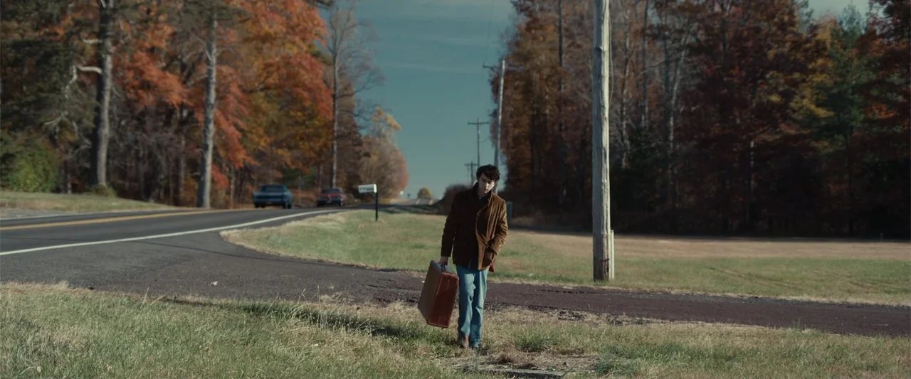 Coming Through the Rye (2016) movie still: fullbody — A rural asphalt road cuts through a landscape featuring autumn foliage and utility poles. A…
