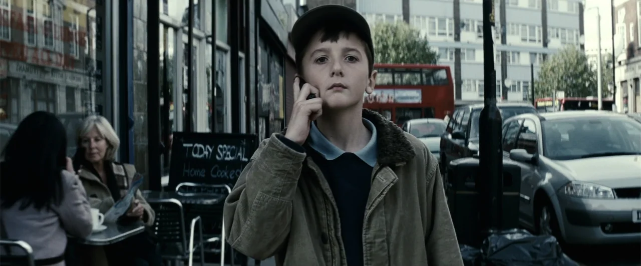 Hereafter (2010) movie still: medium — A city sidewalk beside a bustling street features a cafe with outdoor tables. A young boy stands in the…