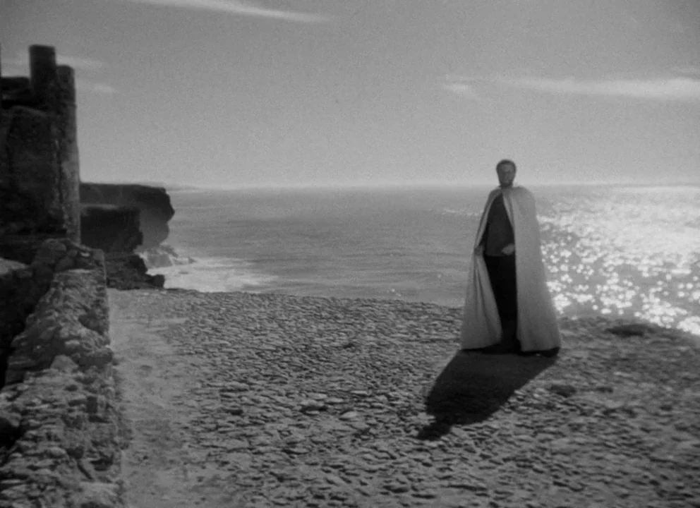 Othello (1952) movie still: wide — A craggy cliff overlooks a vast ocean under a bright sky. A cloaked man stands motionless on the stony ground…
