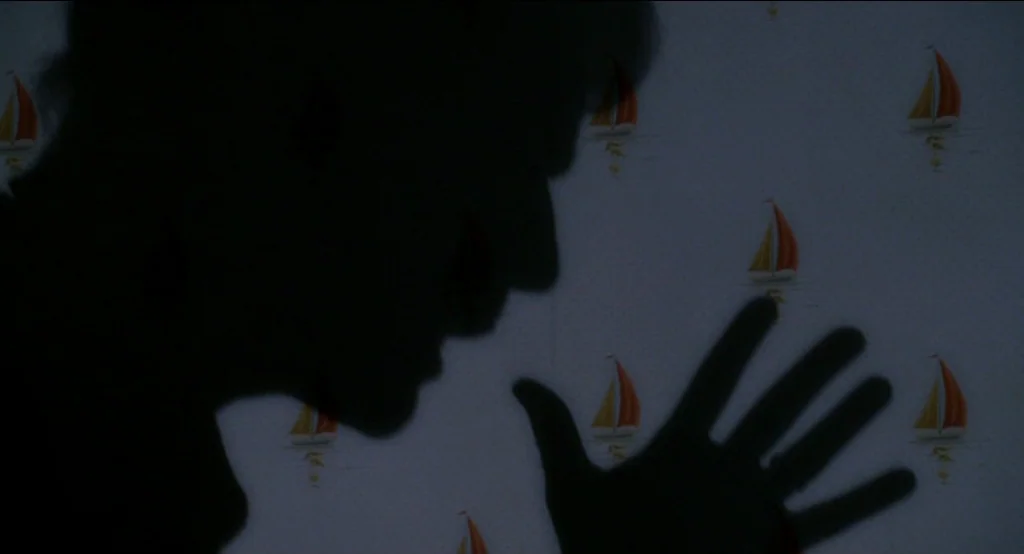 Big Fish (2003) movie still: closeup — A wall covered in wallpaper with a repetitive pattern of small sailboats. A silhouette of a person stands…