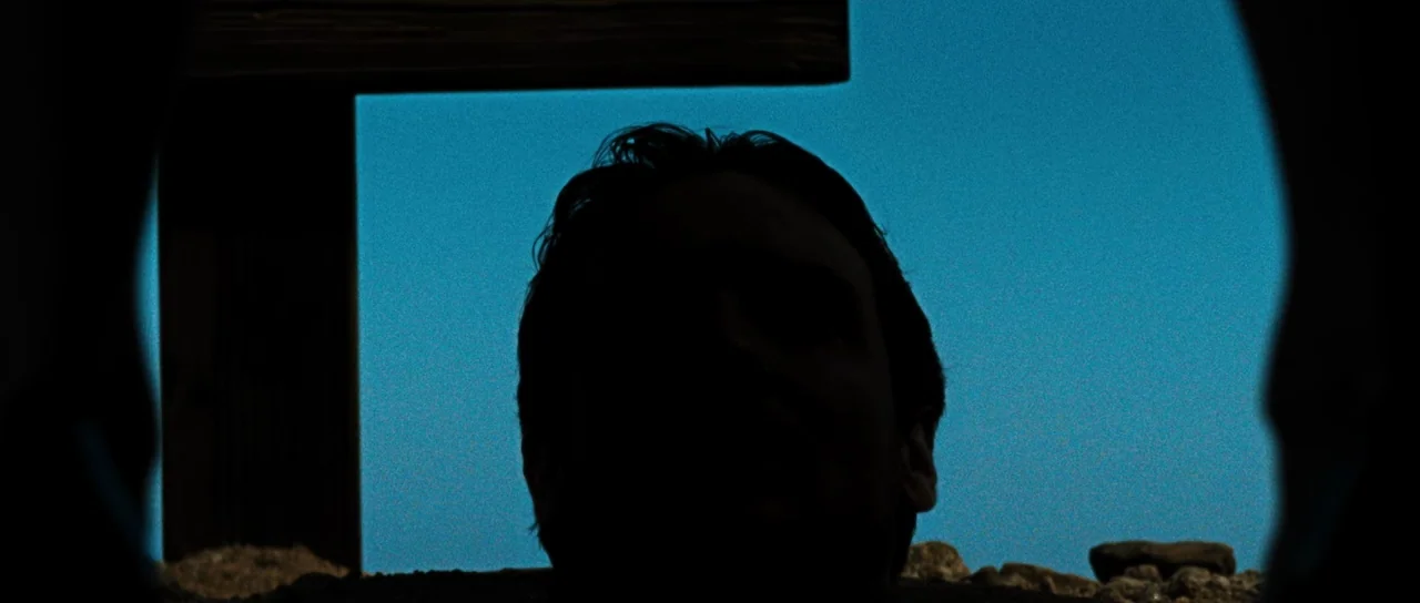 Let The Corpses Tan (2017) movie still: closeup — A man's silhouetted head is centered against a vibrant blue sky. He appears to be positioned…