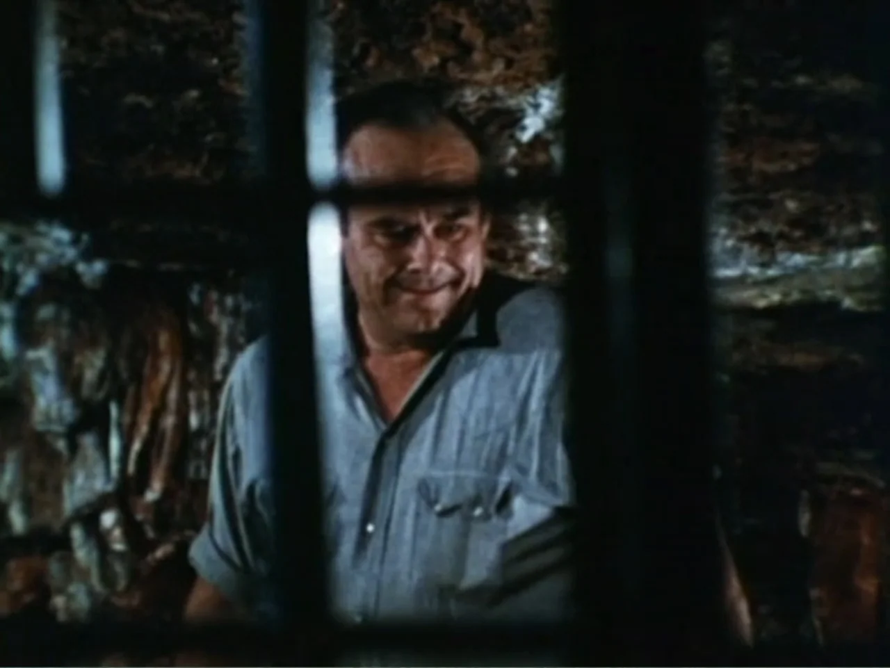 It's Alive (1974) movie still: closeup — A dimly lit stone enclosure with iron bars dividing the foreground and background. A middle-aged man in a…
