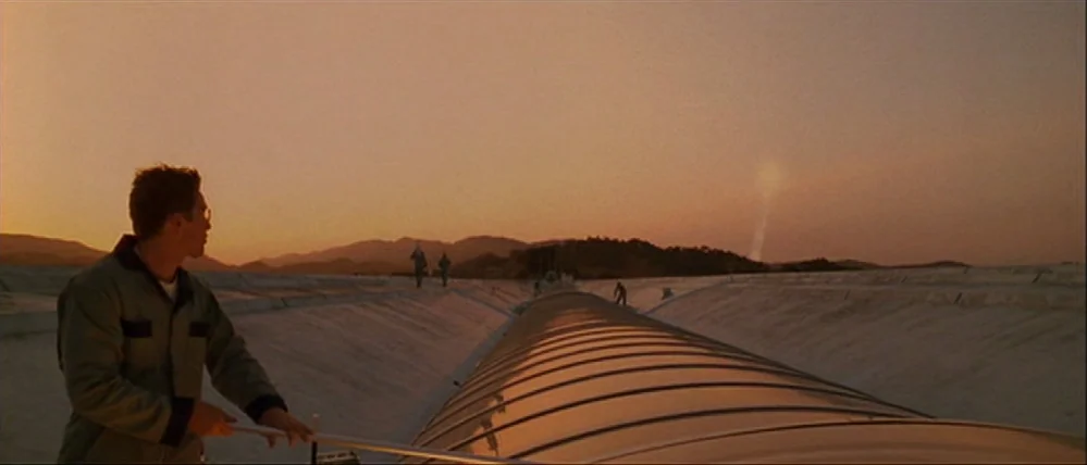 Gattaca (1997) movie still: wide — A vast, concrete-lined water channel spans the landscape under a clear sunset sky. A man stands in the…