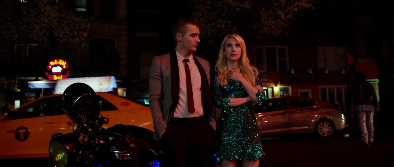 Nerve (2016) movie still: medium — An urban city street at night illuminated by neon signage and street lighting. A young man and woman stand beside…