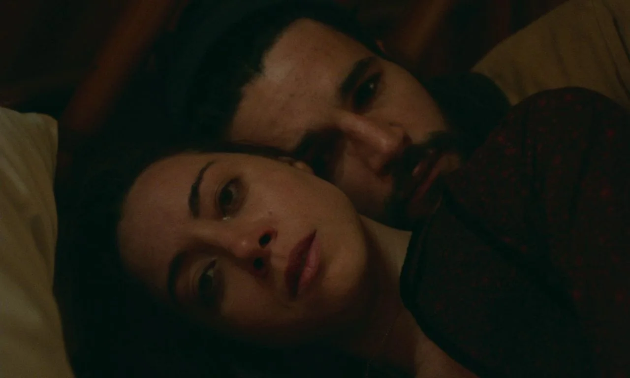 Black Bear (2020) movie still: closeup — A man and woman lie closely together on a surface in dim lighting. They are resting their heads next to…