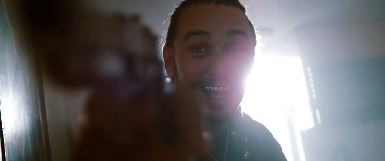 Spring Breakers (2012) movie still: closeup — An interior space is illuminated by a bright, overexposed window in the background. A man with braided…