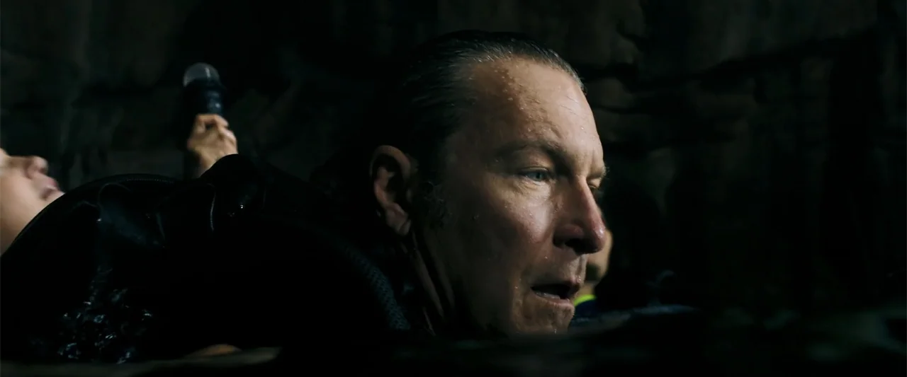 Anaconda (2024) movie still: closeup — A dark, rugged interior space with damp, uneven rock surfaces surrounding the subject. A man in a dark jacket…