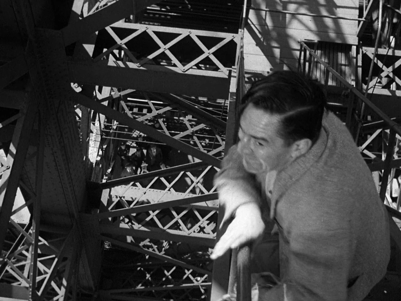 The Naked City (1948) movie still: medium — A man stands high on a metallic lattice tower above a city street. He rests his arms on a steel…
