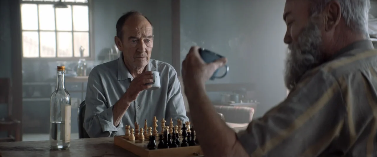 My Neighbor Adolf (2022) movie still: medium — A rustic indoor space features a wooden table with a chess set and glass bottle. Two elderly men sit…