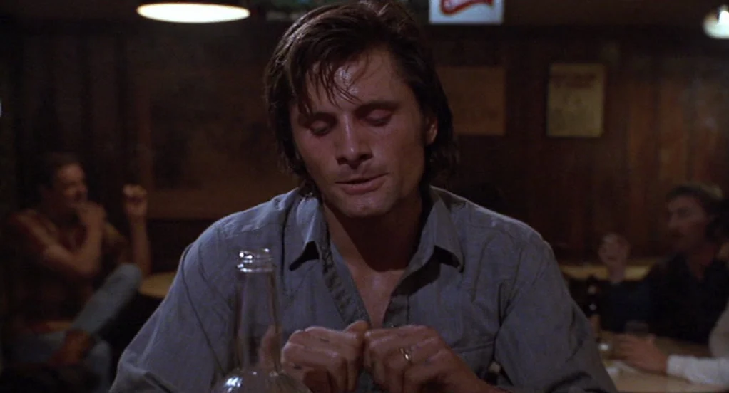 The Indian Runner (1991) movie still: medium — A dark, wood-paneled bar with dim lighting and blurry figures in the background. A man in a blue…