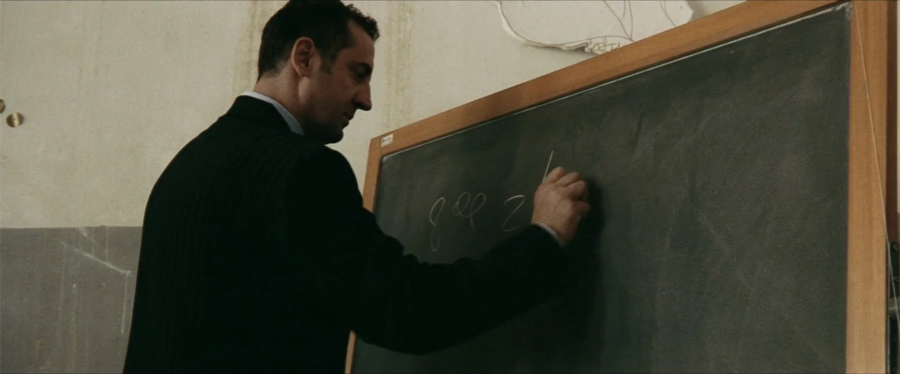 GOMORRAH (2023) movie still: medium — A plain classroom wall features a large, dark green framed chalkboard. A man in a dark suit stands writing…