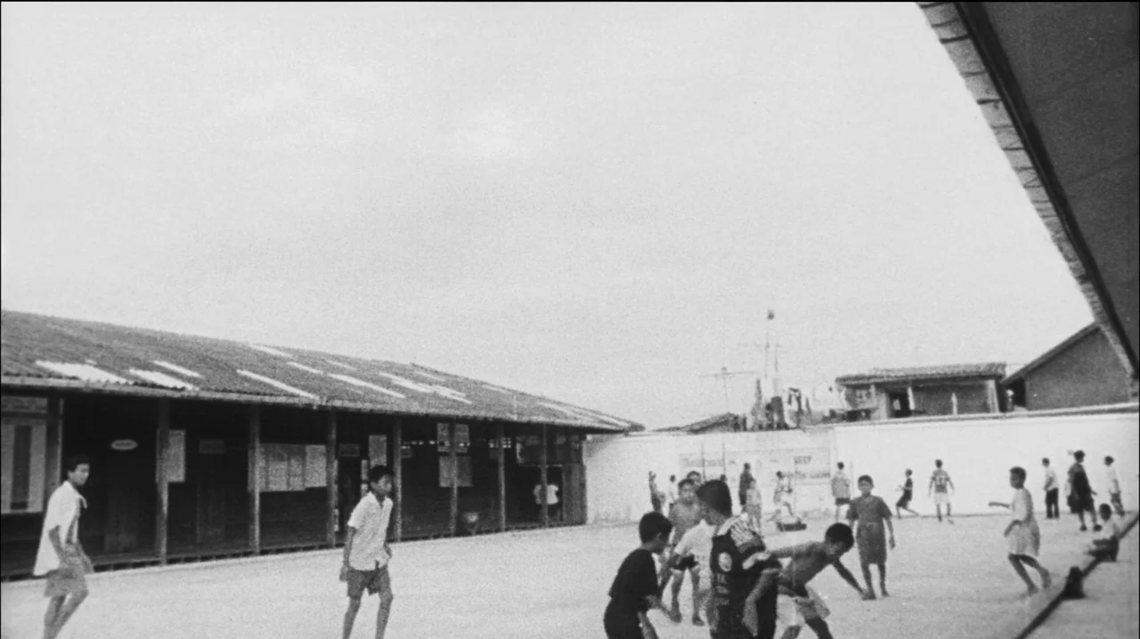 Mysterious Object at Noon (2000) movie still: wide — A wide schoolyard is framed by a low, long building on the left side. Numerous children move…