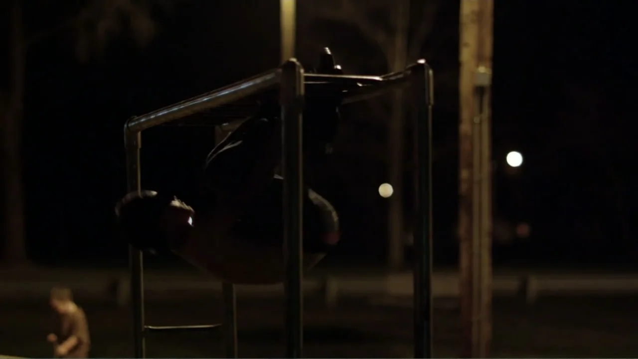 Rich Hill (2014) movie still: medium — A dimly lit outdoor exercise area features a metal monkey bar structure at night. A young man exercises on…