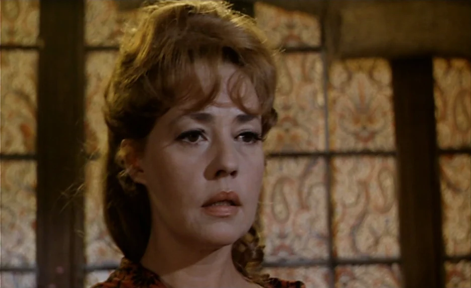 The Immortal Story (1968) movie still: closeup — A woman stands before a wall featuring intricate patterned wallpaper with dark vertical trim. She…